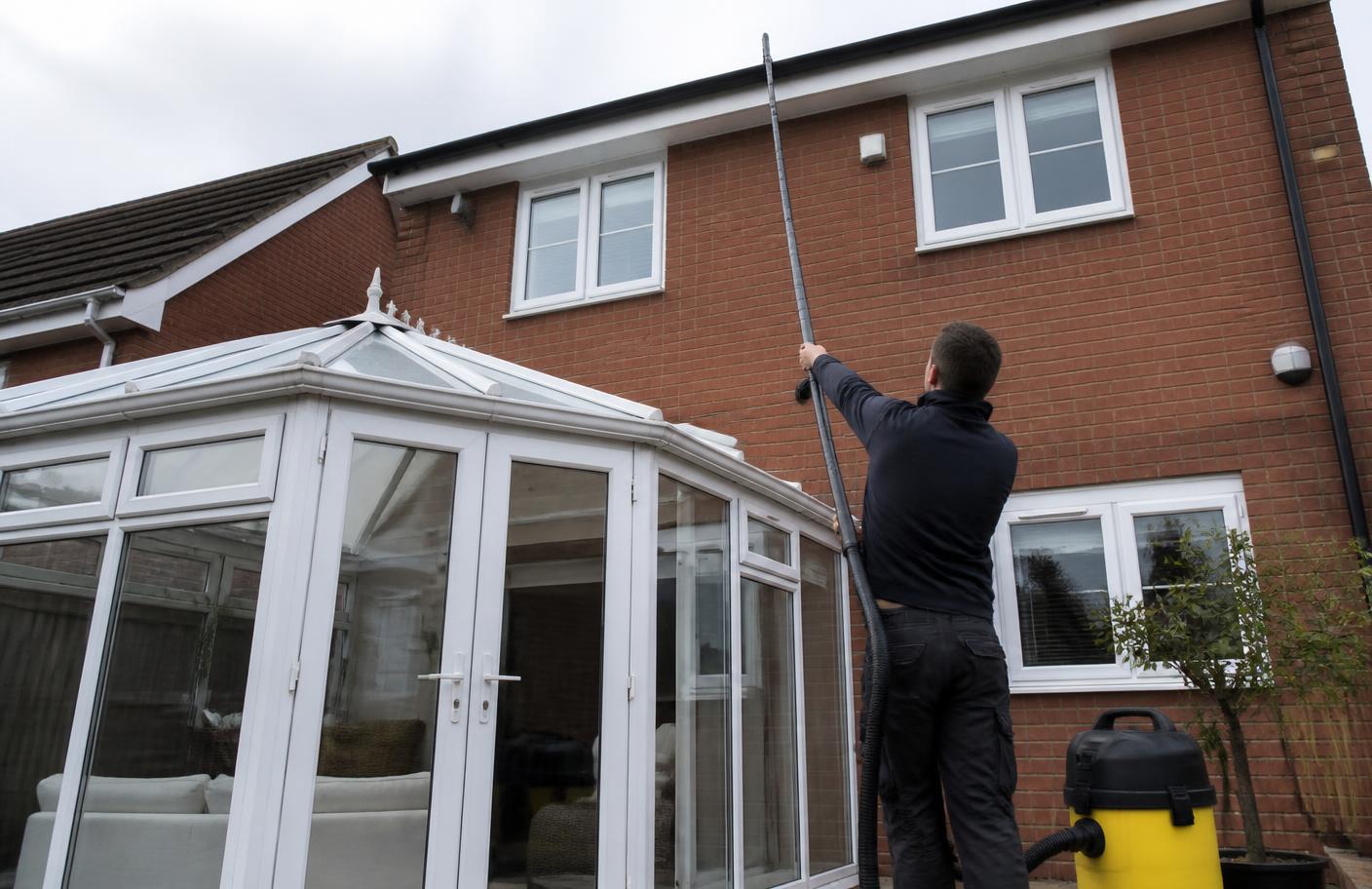 Technician using a ground-operated carbon fibre vacuum pole to clear a gutter above a glass conservatory roof