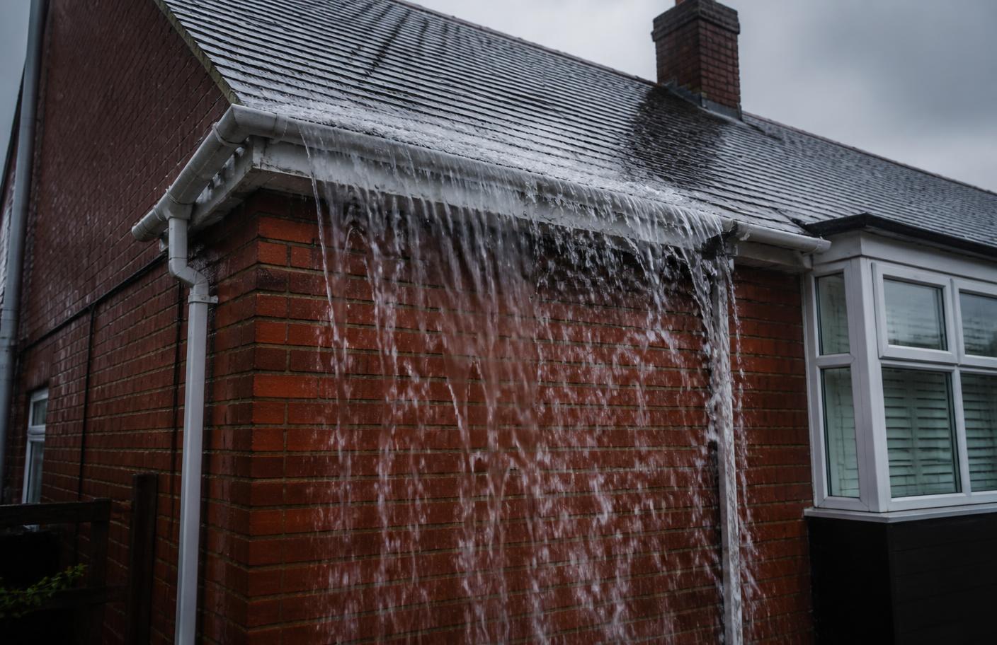 Rainwater cascading over the front edge of a blocked gutter and down the brickwork of a UK semi-detached house during heavy rain